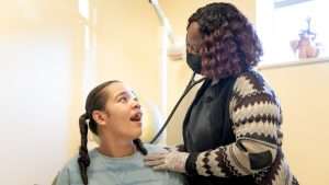 In bright room painted tan, a young girl with black pigtails smiles and looks up at a nurse practitioner as the NP holds a stethoscope to her back at Woods Healthcare's Medical Center.